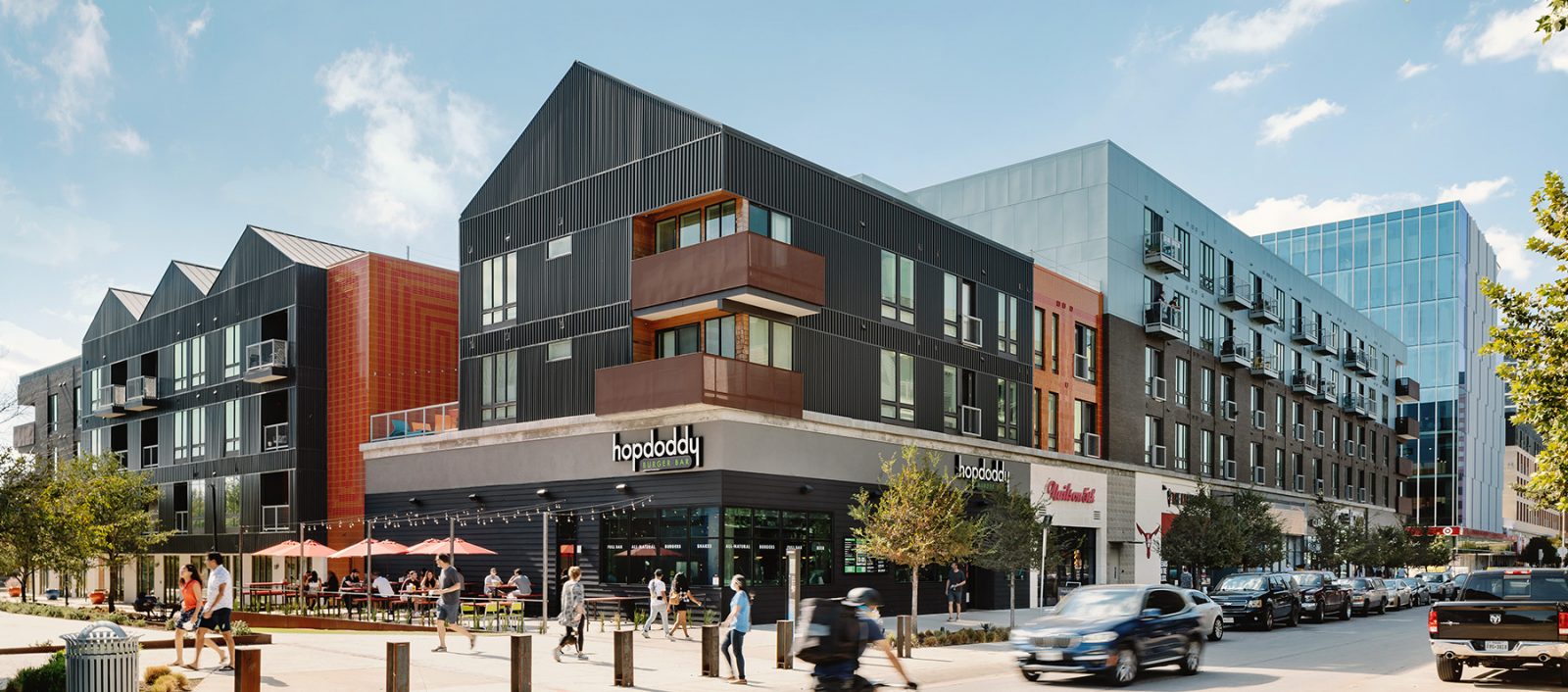 Street view of Plaza Saltillo, featuring modern mixed-use buildings, outdoor dining, and a cyclist passing by.